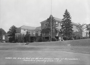 A black and white photograph of the Torresdale-Frankford Country Club clubhouse from a 45 degree angle, viewed from the golf greens. Several trees and shrubbery trim the two-story buildings perimeter. 