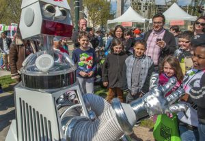 A color photograph of a crowd viewing a humanoid robot outdoors with festival booths and tents in the background