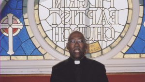 A color photograph of Baba Abiodun in clerical dress in front of a stained glass window