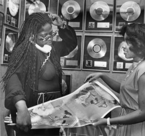 Black and white photograph of two women looking at photos of recording artists, several gold or platinum records hang on the wall behind.