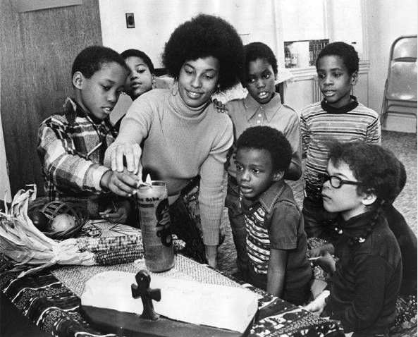 A black and white photograph of a teacher and a group of children lighting a candle with other Kwanzaa symbols around