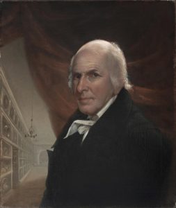 A self-portrait painting by Charles Willson Peale. He stands (depicted bust-up) in front of a red curtain with a hallways behind him to the left. Shelves line the hallway wall, filled with various implied antiques. A delicate chandelier hands about halfway down the hallway above an open door to the left amongst the shelving. Peale is bald on top with brown eyes,wearing a black overcoat over a white-collared shirt.