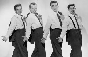 Black and white photograph of four young men in white dress shirts, ties, and dark pants with suit jackets draped over their left shoulders.