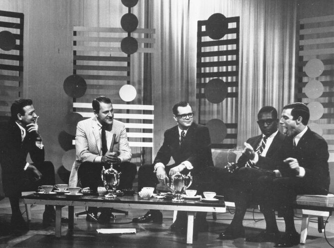 six men sit around a coffee table (with coffee cups and a chromed kettle atop it), all leaning forward in discussion, presumably, about music. the backdrop of the set is decorated with standing wooden slats, like vertical fences and modernist circles. The men all wear suits and ties, most smirking, looking toward Jerry Blavat (far right), who has his arms outstretched and is sticking out his tongue.