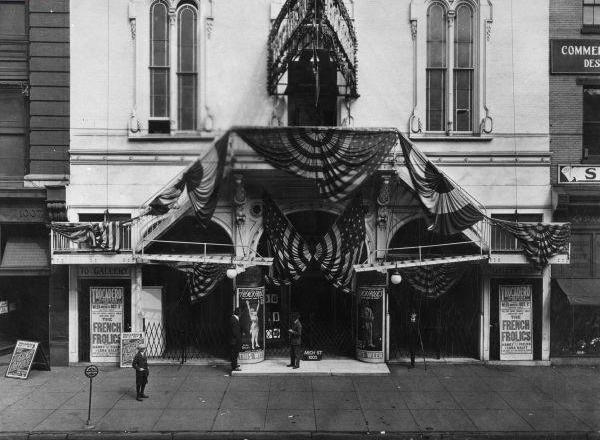 Black and white photograph of the facade of a white building with several banners draped above the entrance. Two signs at either side of the entrance advertise