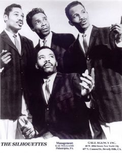 Black and white photograph of four young African American men dressing dark suit jackets, ties, and light pants. Three men are standing while one kneels in front, all are looking off to their left, at least two appear to be singing.