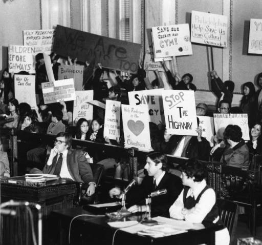Demonstrators to save Chinatown are shown here in 1973, holding up their signs as Lynne M. Abraham from the Redevelopment Authority and James Martin of the Old Philadelphia Development Corporation testify at a hearing at City Hall in Philadelphia.