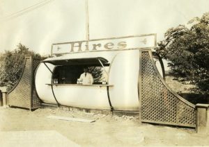 Photograph of a Hires Root Beer stand, situated along the side of a road. The stand is oblong, shaped like a capsule, with one large window in the center for serving customers. Above the stand a sign runs along top and reads "Hires". A clerk stands at the window.