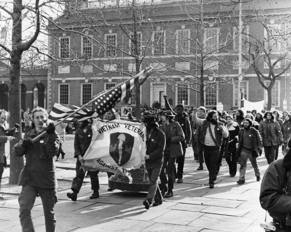 A group of Vietnam Veterans marches by from  right to left, Independence Hall is behind them.  The first one carries an American flag, positioned upside-down on its pole, while two behind him carry a tattered banner that reads