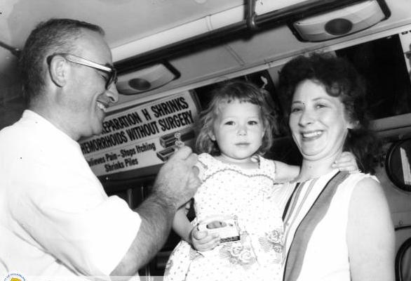 A black and white photo of a doctor administering a vaccine to a young girl being held by her mother. The vaccine is being administered on a city bus used as a mobile clinic.