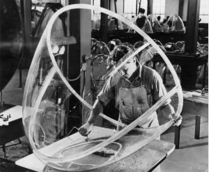 Black and white photograph of a man in overalls holding two clear pieces to form the nose of an airplane together.