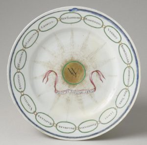 a color photograph of a white dish. The outer rim is decorated with a blue snake biting its tail. Inside this ring is a ring of fifteen chain links with the name of one state in each. In the center of the plate is a gold disk with a monogram and a banner with a latin phrase.