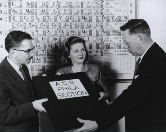 Black and white photograph of two men and a woman. Behind the group, a periodic table hangs on the wall. The man to the left is handing over a plaque that reads "A.C.A Phila. Section" to the man on the right.