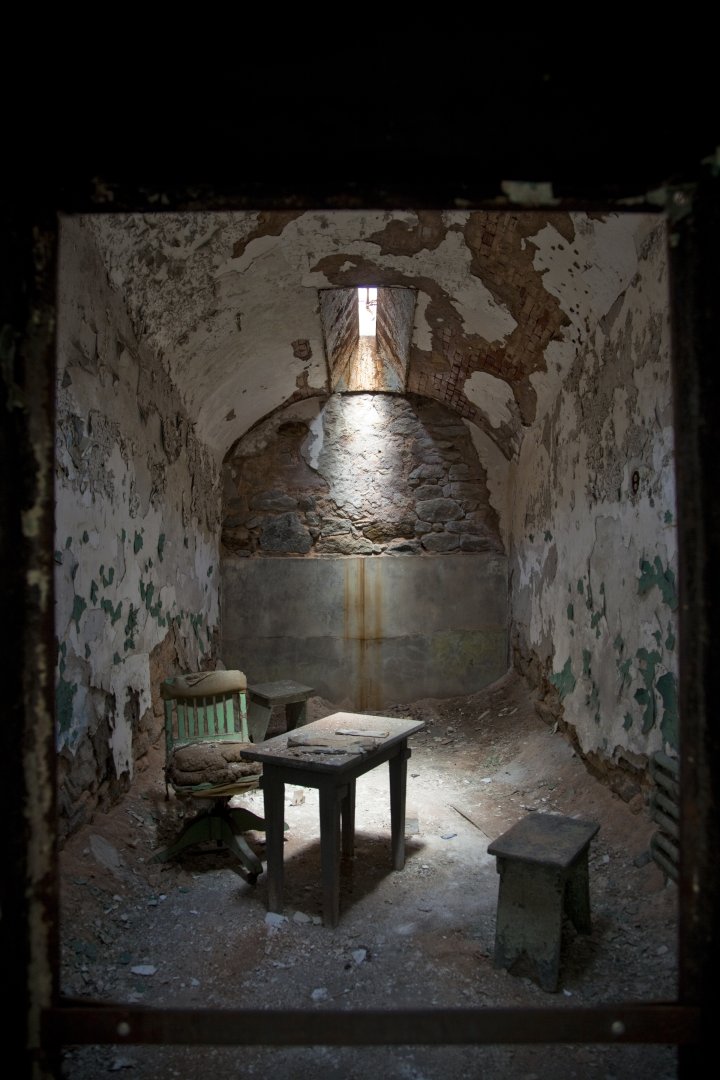 A color photograph of a cell at EAstern State Penitentiary. The walls and ceiling are decaying. There is a narrow skylight in the ceiling allowing light to reach a chair, desk, and two small stools in the cell, all in a state of decay.