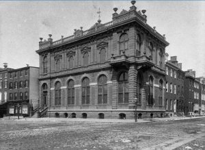  A black and white photograph of the German Society of Pennsylvania, a two-story red brick building with arched windows on the first floor, and prominent balconies and faux Greek columns on on the second floor. Decorative stone railings, urns, and cupolas adorn the roof.