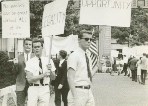 A black and white photograph of picketers whose signs read: opportunity, equality, and no society can be great without All of its citizens