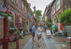 A color photograph of tourists taking a photo on Elfreth's Alley, a narrow cobblestone and brick street lined with restored eighteenth and nineteenth century row homes.