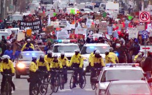 color photo of a line of bike cops in the foreground, demonstrators in the background, moving south on Broad Street on MLK Day 2015.