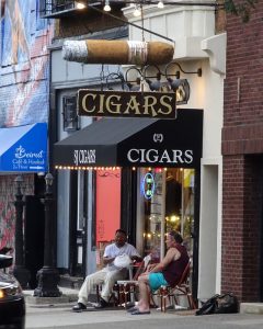 color photo of a cigar shop with two men seated outside on the sidewalk.