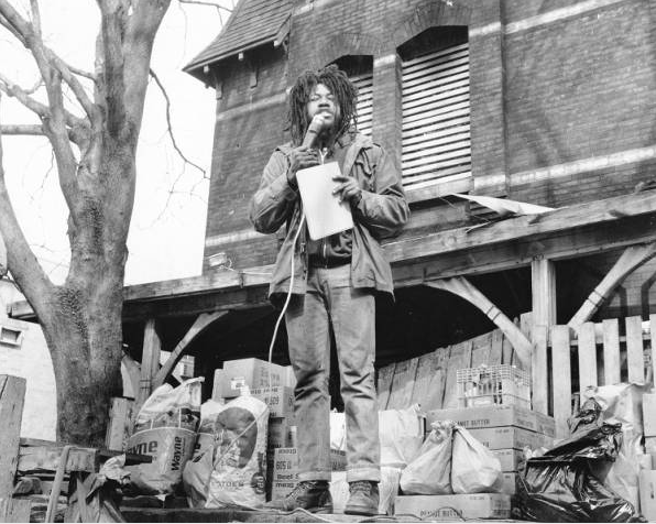 A black and white photograph of a man speaking into a microphone in front of a barricaded home