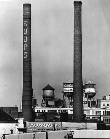 A black and white photograph of two smokestacks and a cluster of water towers. The smokestacks have large letters reading