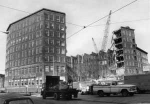 A black and white photograph of a multi-story factory complex being demolished