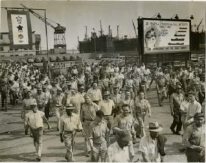 A photograph of a crowd of a mixed group of black and white men in work clothes leaving Sun Ship's complex in Chester circa 1950. Behind them a sign reads "30 years of progressive activity"