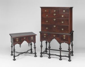 A color photograph of a wooden chest of drawers and dressing table with decorative carvings, thin legs, and brass fittings.