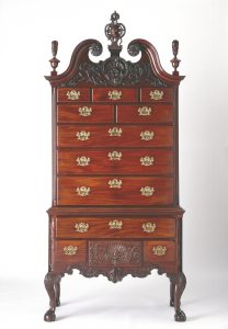 A color photograph of a wooden chest of drawers with elaborate carvings on the header and base, and brass fittings.