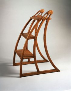 A color photograph of a curved wooden step ladder. The tops of the hand rails are carved into a stylized donkey and elephant.