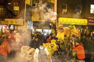 a color photograph of a lion dance in Chinatown showing dancers wielding large chinese lion puppets and smoke clouds from firecrackers