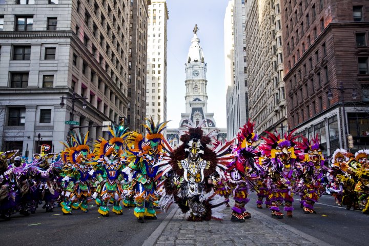 A photograph of mummers -- men dressed in elaborate, colorful costumes -- walking down Broad Street with City Hall in the background