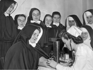 a black and white photograph of a nurse drawing blood from a nun while several other nuns and postulants look on