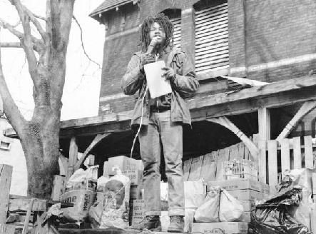 a black and white photograph of Delbert Africa speaking into a microphone in front of his home, with a pile of foodstuffs around his feet