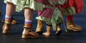 close up photograph of mummer's feet, from the shin down. Pieces of colorful costumes flow over work boots that have been spray painted gold