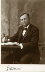 a black and white photograph of John Wanamaker in a suit seated at a desk. He faces the camera, and in his right hand holds a pen as if writing.