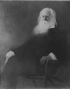 a black and white photograph of Walt Whitman as an elderly man. He sits in a chair with legs crossed.