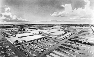 an artists' conception of the Cherry Hill Mall. It is drawn from an aerial perspective. In the center is a cluster of large, flat white buildings. The buildings are surrounded by extensive surface parking lots full of parked cars.