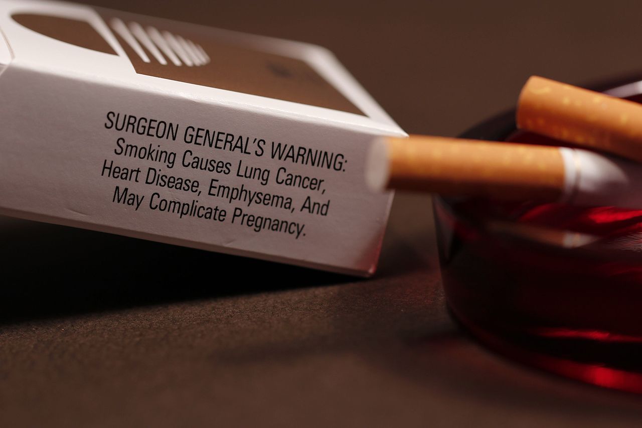 a color photograph of a pack of cigarettes focusing on the Surgeon General warning. An ashtray with two cigarette butts is in the foreground.