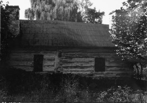 A black and white photograph of a small log cabin with wood shingle roof and stone chimney