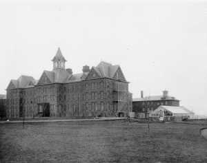A black and white photograph of the main building of Delaware State Hospital at Farnhurst with grounds and greenhouse in the background