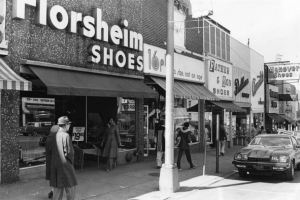 A black and white photograph of shoppers walking through a busy business district. Sign on shop reads Florsheim Shoes. 