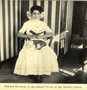 A black and white photograph of Deborah Kallikak reading a book with a tabby cat on her lap. Text reads "Deborah Kallikak, as she appears to-day at the Training School"