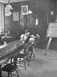 A black and white photograph of a room in the Elwyn Training School. A nurse sits with a small child on her lap. The child holds a small tuba-like musical instrument. In the background is a chalk board on an easel with musical notation hand written on it.