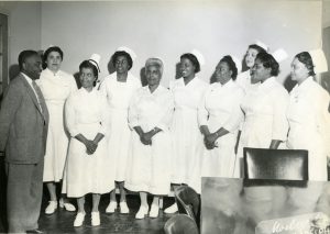 a black and white photograph of nine African-American women in white nursing uniforms and caps. An African-American man in a suit stands with them.