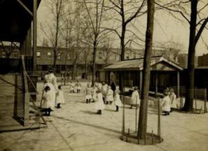 Small children in white uniforms play outside as nurses stand watching