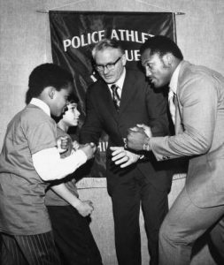 Joe Frazier poses with his fist raised opposite of PAL kids. In the background of the photograph is Police Commissioner Joseph O'Neill.