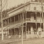 A nineteenth century photograph of the Maple Spring Hotel. The building is rectangular in shape with a pronounced porch along its exterior. Behind the building is a forest.