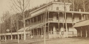 A nineteenth century photograph of the Maple Spring Hotel. The building is rectangular in shape with a pronounced porch along its exterior. Behind the building is a forest.