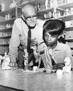 A black and white photograph of a Police Athletic League officer standing over the shoulder of a teenage woman, as she paints a ceramic sculpture.